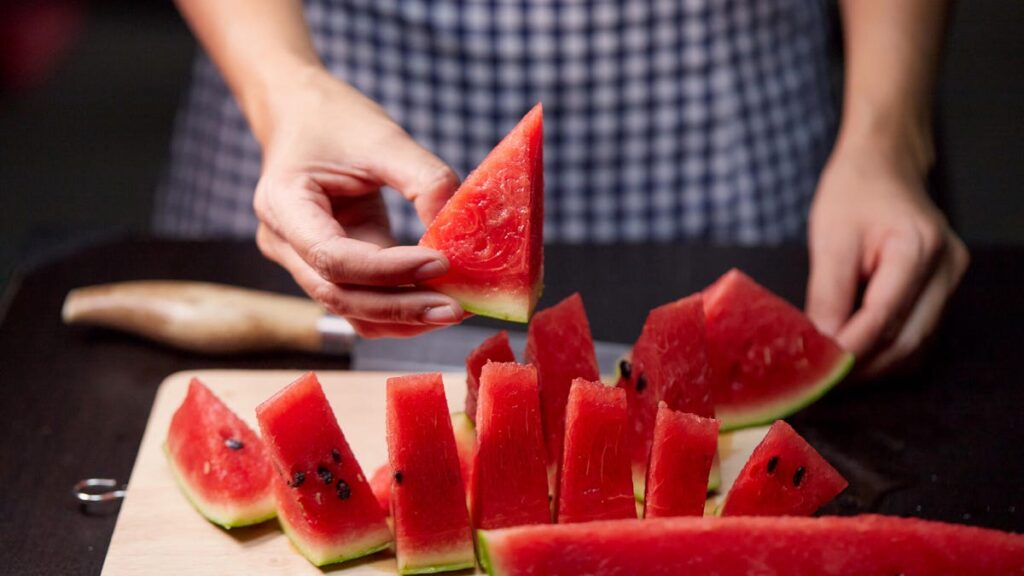A close up of a person wearing blue gingham cutting up watermelon on a wood cutting board.