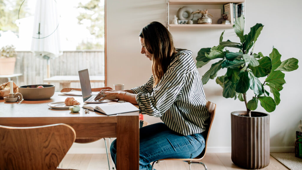 woman at a laptop at a kitchen table with a plant behind her