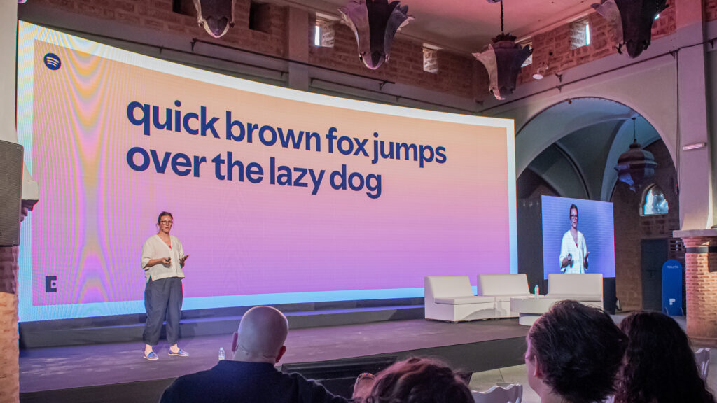 'quick brown fox jumps over the lazy dog' on the screen with a woman talking on a stage