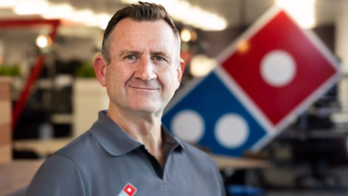 Domino’s Pizza parts ways with chief who bet on fried chicken Andrew Rennie stands indoors in a Domino’s uniform, with a large Domino’s Pizza logo visible in the background.