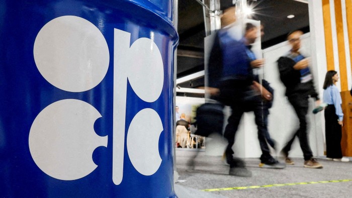 Opec+ to pause oil output rises next year after warnings of glut A large blue barrel with the Opec logo is displayed in the foreground as people walk past at a conference venue.