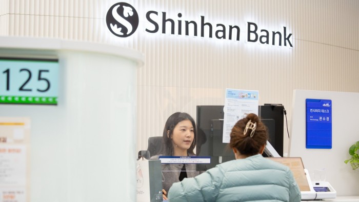 South Korean banks take the lead to bridge gender inequalities A Korean woman sitting at a counter behind a glass screen serving another woman below a sign saying ‘Shinhan Bank’