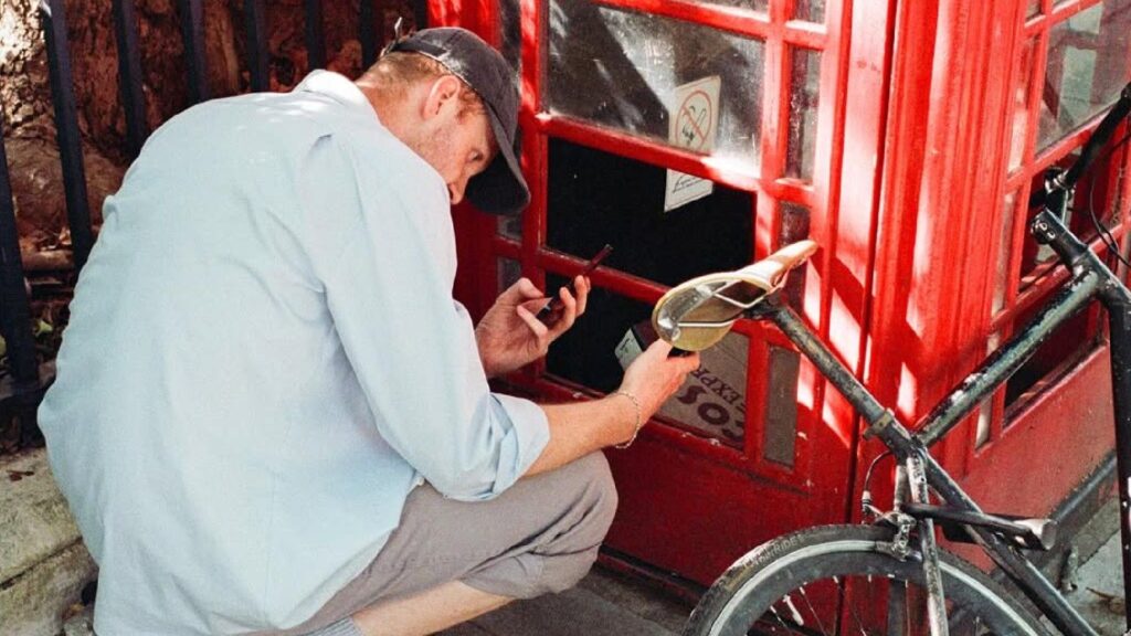 This guerrilla network sharing art gallery memberships is my new favourite thing A man wearing a light blue shirt and a baseball cap is crouching down next to a red telephone booth, appearing to be examining something inside or on the ground while holding a small black object and a bottle.